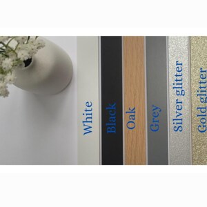 K&ouml;nnte beinhalten: Eine Auswahl an farbigen Bilderrahmenmustern. Die Muster umfassen Wei&szlig;, Schwarz, Eiche, Grau, Silberglitter und Goldglitter. Jedes Muster hat eine Beschriftung mit seinem Farbnamen in blauem Text. Eine wei&szlig;e Vase mit wei&szlig;en Blumen befindet sich in der oberen linken Ecke.
