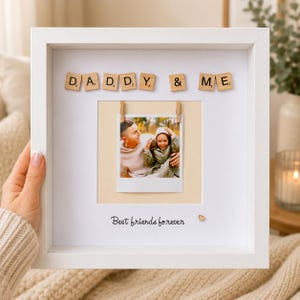 May include: A white square shadow box frame containing a photograph of two people clipped to a cream-colored background. The text DADDY & ME is spelled out in wooden letter tiles above the photo, and the phrase Best friends forever is printed below.