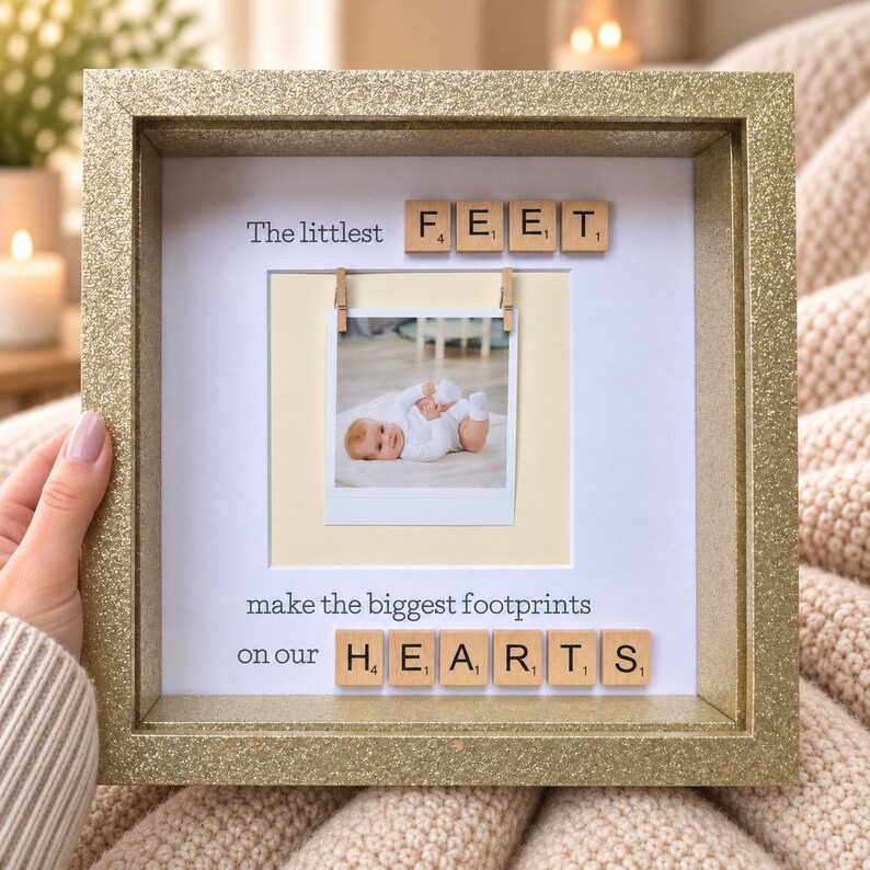 May include: A square gold glitter frame containing a white mat with the text "The littlest FEET make the biggest footprints on our HEARTS". A square photograph of a baby is clipped to the center with two small wooden clothespins.