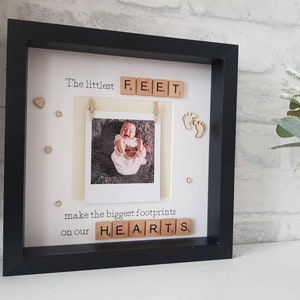 K&ouml;nnte beinhalten: Schwarzer Bilderrahmen mit Babyfoto und Text "The littlest FEET make the biggest footprints on our HEARTS". Das Foto wird von einer kleinen Holzklammer gehalten. Holzherz- und Fu&szlig;abdruck-Akzente sind ebenfalls enthalten.