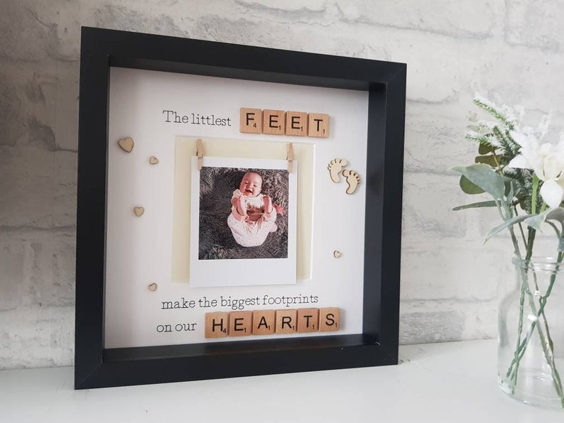 K&ouml;nnte beinhalten: Schwarzer Bilderrahmen mit Babyfoto und Text "The littlest FEET make the biggest footprints on our HEARTS". Das Foto wird von einer kleinen Holzklammer gehalten. Holzherz- und Fu&szlig;abdruck-Akzente sind ebenfalls enthalten.