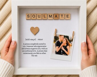 May include: A white square shadow box frame containing wooden letter tiles spelling SOULMATE, a small wooden heart, a printed definition of the word, and a photograph of a couple clipped to the backing with two small wooden clothespins.