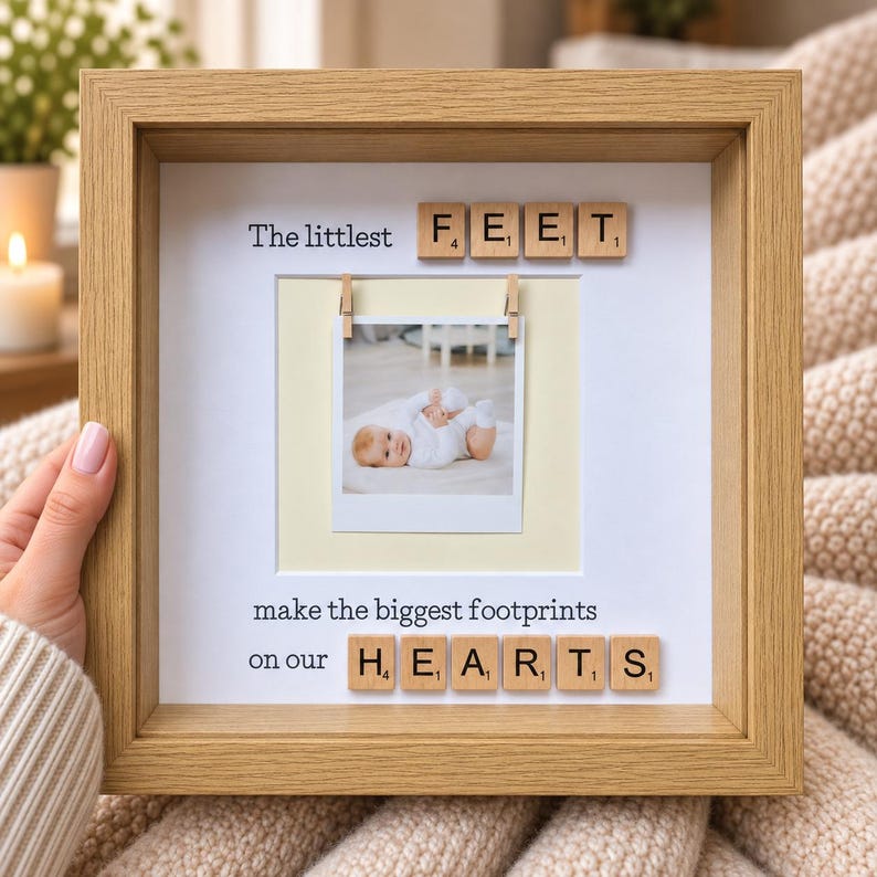 May include: A square light wood shadow box frame containing a white mat with the text The littlest FEET make the biggest footprints on our HEARTS. A small square photograph of a baby is clipped to a string in the center of the frame.