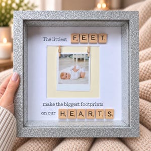 May include: A square silver glitter frame containing a white mat with a central photograph of a baby. The text reads The littlest FEET make the biggest footprints on our HEARTS, with wooden letter tiles spelling FEET and HEARTS.