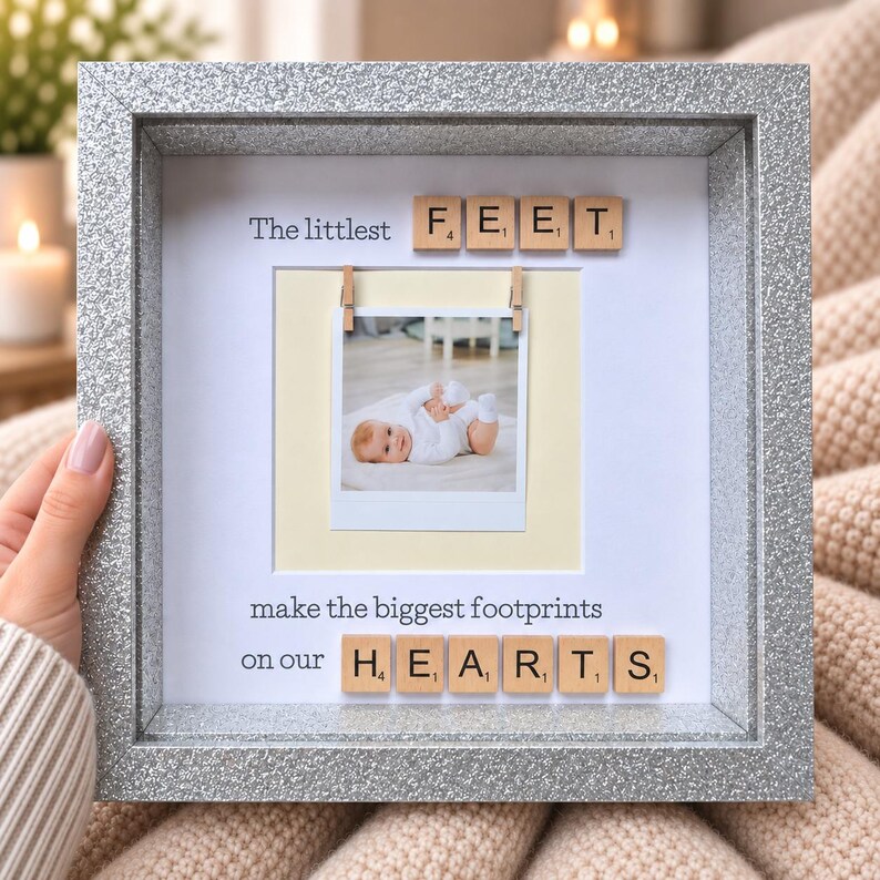 May include: A square silver glitter frame containing a white mat with a central photograph of a baby. The text reads The littlest FEET make the biggest footprints on our HEARTS, with wooden letter tiles spelling FEET and HEARTS.