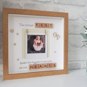 K&ouml;nnte beinhalten: Ein gerahmtes Babyfoto mit dem Text "The littlest FEET make the biggest footprints on our HEARTS". Der Rahmen ist hellbraun, mit wei&szlig;em Hintergrund und kleinen Holzherzen. Ein Babyfoto ist am Rahmen befestigt.