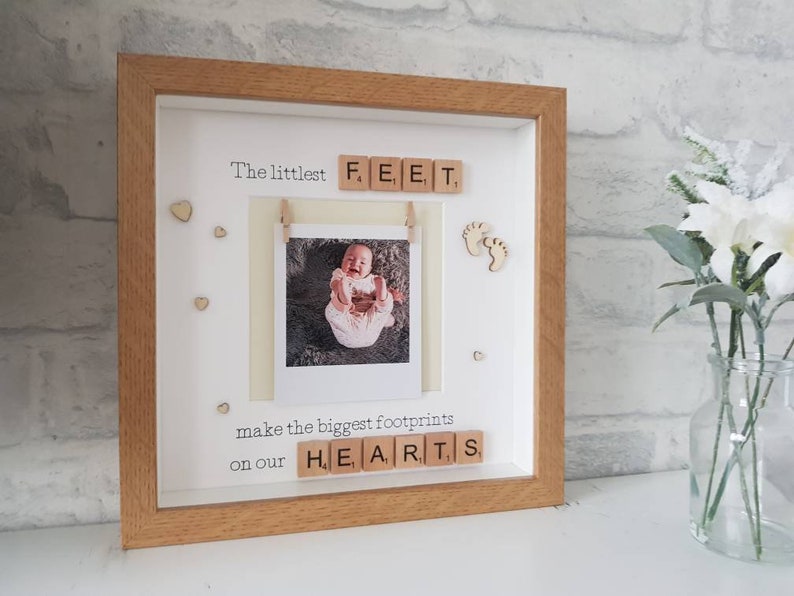 K&ouml;nnte beinhalten: Ein gerahmtes Babyfoto mit dem Text "The littlest FEET make the biggest footprints on our HEARTS". Der Rahmen ist hellbraun, mit wei&szlig;em Hintergrund und kleinen Holzherzen. Ein Babyfoto ist am Rahmen befestigt.