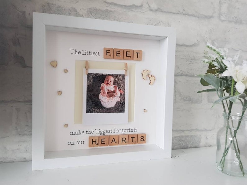K&ouml;nnte beinhalten: Ein wei&szlig; gerahmtes Babyfoto-Display mit dem Text "The littlest FEET make the biggest footprints on our HEARTS." Das Display enth&auml;lt ein Babyfoto, Fu&szlig;-Ausschnitte aus Holz und kleine Herz-Akzente.