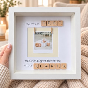 May include: A white square shadow box frame containing a central photograph of a baby. The text reads The littlest FEET make the biggest footprints on our HEARTS, with the words FEET and HEARTS formed by wooden letter tiles attached to the backing.