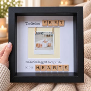 May include: A black square shadow box frame containing a white mat with a central square photo of a baby. The text reads The littlest FEET make the biggest footprints on our HEARTS, with wooden letter tiles spelling FEET and HEARTS.