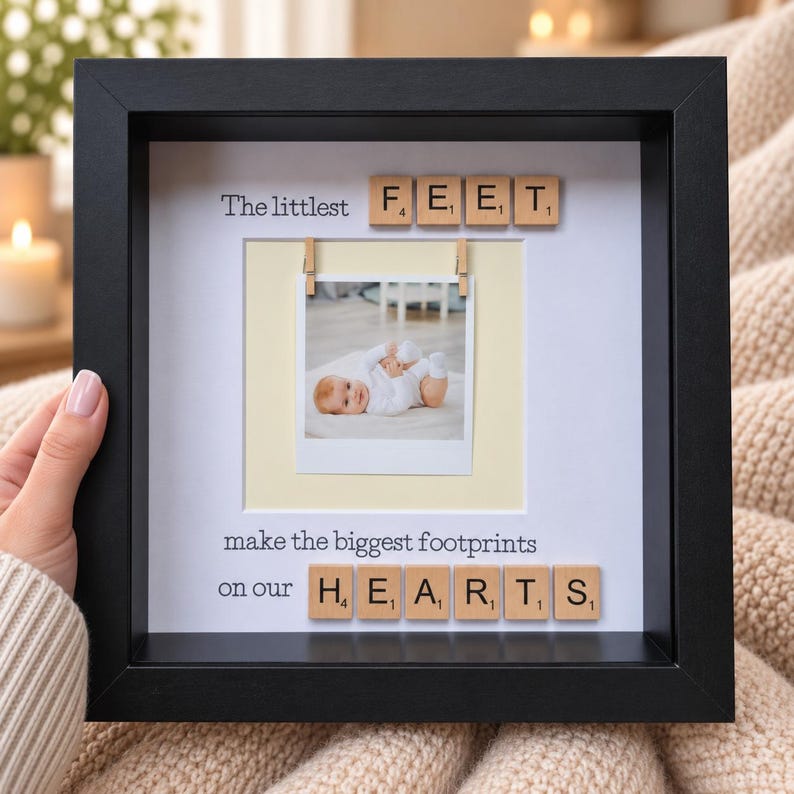 May include: A black square shadow box frame containing a white mat with a central square photo of a baby. The text reads The littlest FEET make the biggest footprints on our HEARTS, with wooden letter tiles spelling FEET and HEARTS.