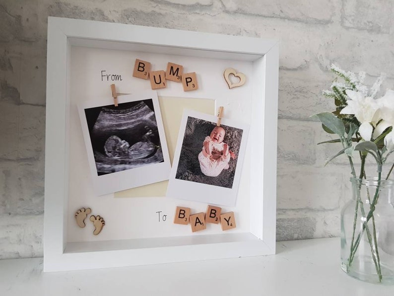 May include: A white picture frame with a white mat. The frame has two photos inside, one of an ultrasound image and one of a baby. The photos are held in place with small wooden pegs. The frame also has a wooden heart, wooden baby feet, and wooden letter tiles that spell out 'From Bump to Baby'.