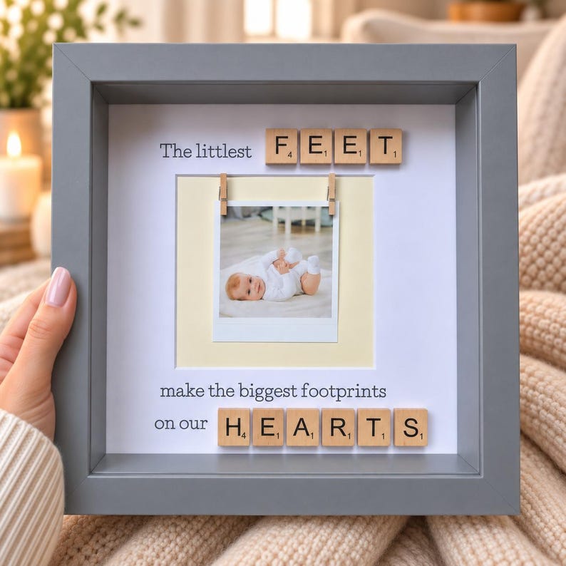 May include: A square gray shadow box frame containing a white mat with the text The littlest FEET make the biggest footprints on our HEARTS. Wooden letter tiles spell out FEET and HEARTS. A square photo of a baby is clipped to the center.