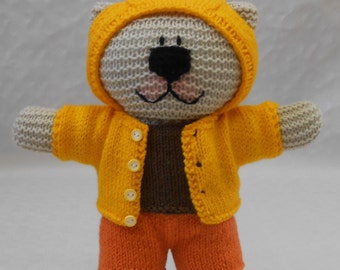 May include: A knitted grey teddy bear wearing a yellow hooded jacket and orange shorts. The bear has a black nose and pink cheeks.