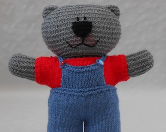 May include: A grey knitted teddy bear wearing a red shirt and blue dungarees. The bear has a black nose and a friendly expression.