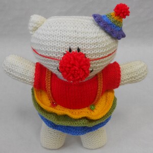 Könnte beinhalten: Ein weißes, gestricktes Stofftier, das ein rotes, gelbes, grünes und blaues Clownskostüm mit einem lila und regenbogenfarbenen Hut trägt. Das Stofftier hat eine rote Pomponnase.