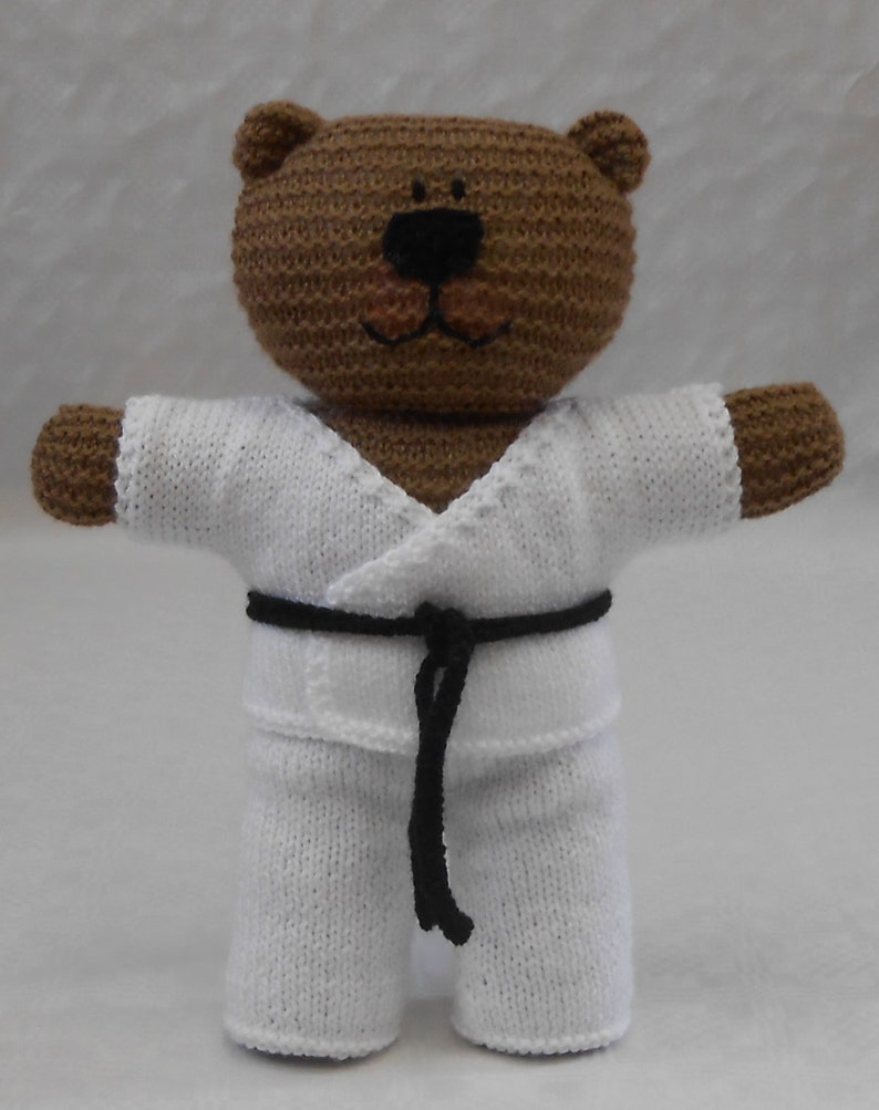 May include: A brown knitted teddy bear wearing a white karate uniform with a black belt.