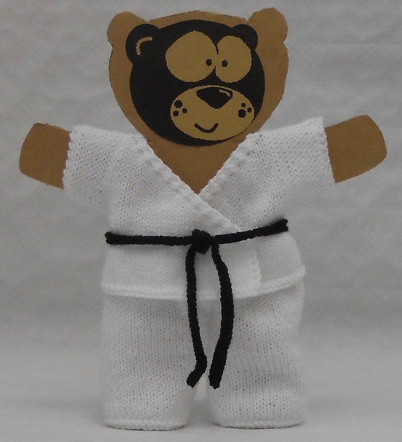 May include: A white knitted karate uniform with a black belt, worn by a brown teddy bear with a black bear face.