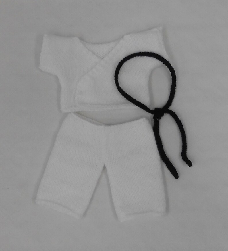 May include: White knitted doll clothes set with a black belt. The set includes a short-sleeved top with a wrap-around closure and a pair of shorts.