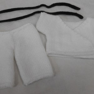 May include: White knitted baby outfit with a long-sleeved shirt and trousers. The shirt has a wrap-style fastening. The outfit is made of soft, white yarn.