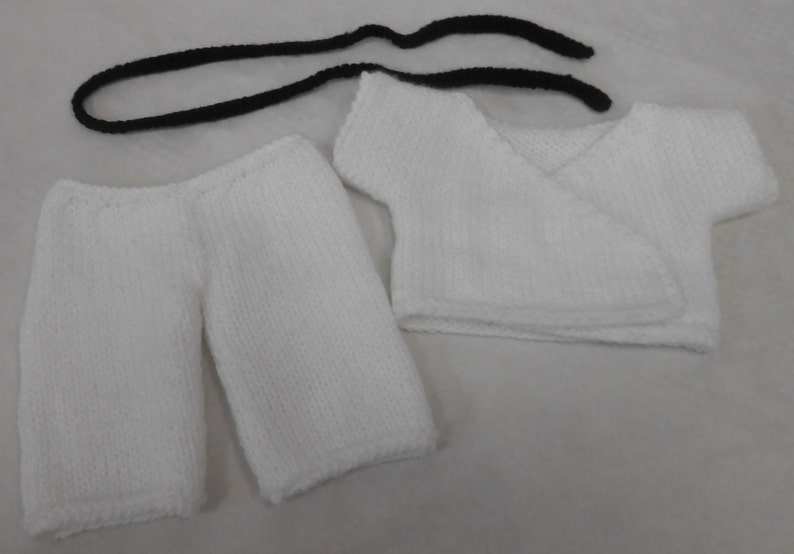 May include: White knitted baby outfit with a long-sleeved shirt and trousers. The shirt has a wrap-style fastening. The outfit is made of soft, white yarn.
