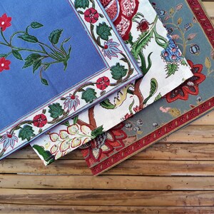 May include: Three fabric napkins with floral patterns. The top napkin is blue with a white border and red flowers. The middle napkin is white with a blue border and red, yellow, and green flowers. The bottom napkin is green with a red border and red flowers.