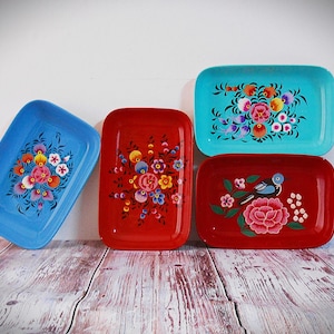 Könnte beinhalten: Vier rechteckige Tabletts mit handgemalten Blumendesigns. Die Tabletts sind in Blau- und Rottönen gehalten, mit bunten Blumen und einem Vogel. Die Tabletts werden auf einer Holzoberfläche präsentiert.