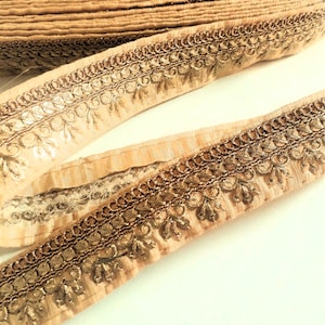 May include: Close-up of a roll of beige fabric trim with intricate gold-colored embroidery along the edges. The trim features a repeating floral pattern and is likely used for embellishing clothing or crafts.