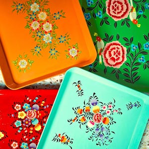 Pode incluir: Quatro tabuleiros retangulares de metal nas cores laranja, verde, vermelho e azul-claro. Cada tabuleiro apresenta motivos florais pintados à mão com pétalas, folhas e caules intrincados. O tabuleiro verde inclui ilustrações de aves.