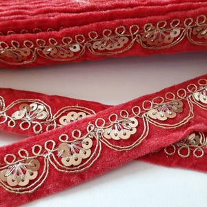 Puede incluir: Borde de terciopelo rojo con lentejuelas y bordados dorados. El borde presenta un diseño festoneado con un patrón repetitivo de lentejuelas y bordados.