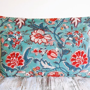 Peut inclure: Coussin rectangulaire avec un fond turquoise et un motif floral rouge, blanc et bleu. Le motif présente de grandes fleurs, des feuilles et des vignes. Le coussin est posé sur une surface en bois clair.