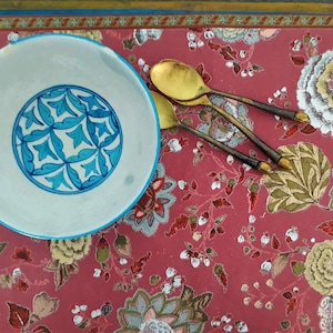May include: A white ceramic bowl with a blue and white floral design sits on a red floral patterned tablecloth with two gold spoons.