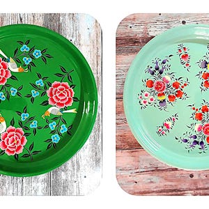 Peut inclure: Deux plateaux ronds en métal avec des motifs floraux peints. Le plateau de gauche est vert avec des oiseaux blancs et des fleurs roses. Le plateau de droite est bleu clair avec des fleurs rouges et violettes.