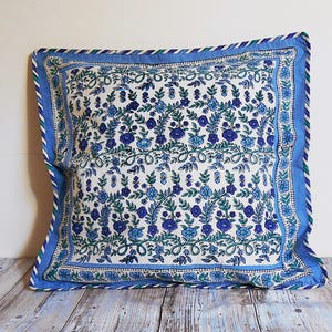Peut inclure: Coussin carré avec un fond blanc et un motif floral bleu. Le coussin a une bordure bleue avec une garniture rayée. Le motif floral présente différentes nuances de bleu et de vert, créant un accent décoratif pour la maison.