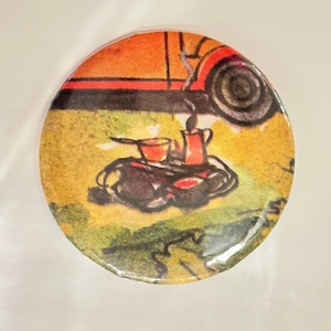 May include: A round, colorful button with a vintage illustration of a red car parked on a grassy field. The car is partially visible, and the focus is on a small camping scene with a kettle, cups, and a small fire.