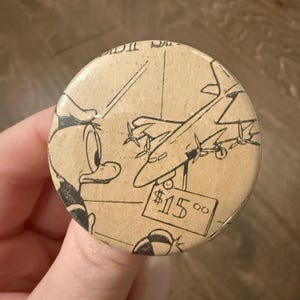 May include: A round button featuring a vintage cartoon illustration of Donald Duck looking at a biplane. The button has a tan background with black line art and the price tag reads $15.00.