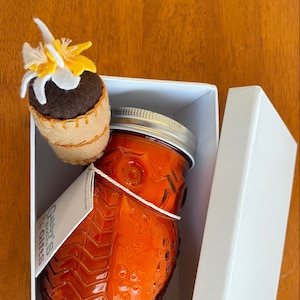 Puede incluir: Una caja de regalo blanca con la tapa abierta, que revela un frasco de vidrio de salsa naranja con una tapa de fieltro marrón en forma de búho. El frasco tiene una etiqueta blanca con el texto "Gabby's Garden & Gifts". Una pequeña flor de fieltro marrón con pétalos amarillos está sentada encima de la caja.