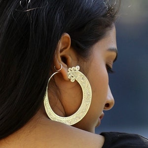 Puede incluir: Una mujer con el pelo largo y negro que lleva una camisa negra y grandes pendientes de aro de plata.