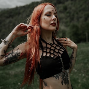 Peut inclure: Une femme aux longs cheveux roux portant un haut noir à bretelles avec un motif en maille. Elle a plusieurs tatouages sur les bras et le torse, et porte un collier et des boucles d'oreilles en argent. Elle se tient dans un champ avec de l'herbe verte et un ciel nuageux en arrière-plan.