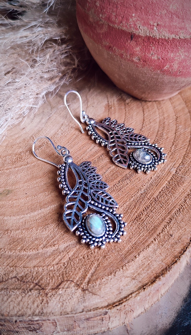 May include: Silver-toned dangle earrings with a leaf design and a central oval-shaped stone. The earrings have a detailed, openwork pattern and a hook closure. The background is a wooden surface with a blurred terracotta pot.