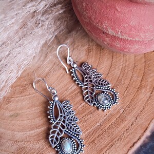 May include: A pair of silver-colored dangle earrings with a leaf design. Each earring features a central oval-shaped stone and intricate detailing. The earrings are displayed on a wooden surface, with a terracotta pot in the background.