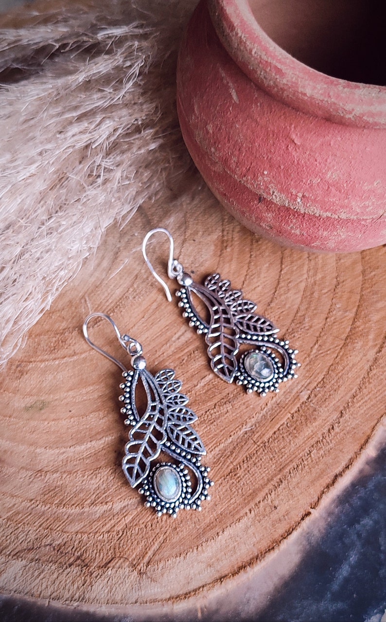 May include: A pair of silver-colored dangle earrings with a leaf design. Each earring features a central oval-shaped stone and intricate detailing. The earrings are displayed on a wooden surface, with a terracotta pot in the background.