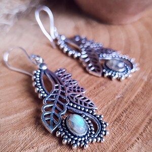 May include: Silver-toned dangle earrings with a leaf design and a central, oval-shaped stone. The earrings feature intricate detailing, including openwork leaves and a beaded border. The background is a wooden surface.