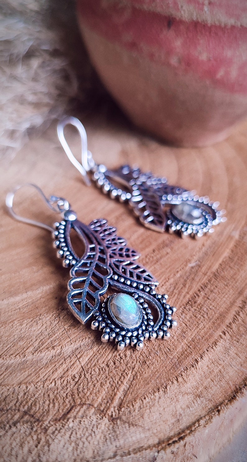 May include: Silver-toned dangle earrings with a leaf design and a central, oval-shaped stone. The earrings feature intricate detailing, including openwork leaves and a beaded border. The background is a wooden surface.
