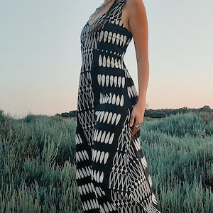 Peut inclure: Une robe longue noire sans manches avec un motif de goutte d'eau blanc. La robe a un col en V et une jupe fluide. Le modèle se tient dans un champ de hautes herbes. La robe est de style bohème.