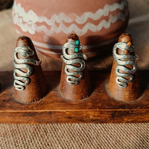 Può includere: Tre anelli a forma di serpente argentati esposti su supporti in legno. Gli anelli presentano dettagliati disegni di serpenti, uno con accenti turchesi. Sono presentati su un vassoio di legno, con un vaso di ceramica decorativo sullo sfondo.