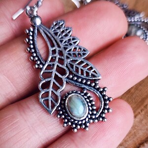 May include: Silver-toned leaf-shaped earrings with intricate cut-out details and a small, oval, iridescent stone. The earrings have a hook closure and are displayed on a hand, showcasing the delicate design and craftsmanship. The earrings are a piece of jewelry.