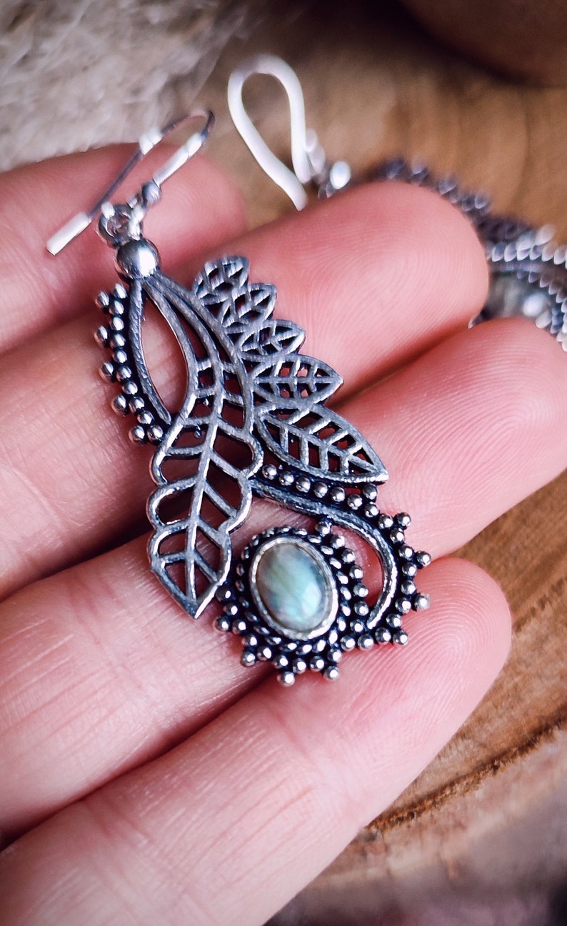 May include: Silver-toned leaf-shaped earrings with intricate cut-out details and a small, oval, iridescent stone. The earrings have a hook closure and are displayed on a hand, showcasing the delicate design and craftsmanship. The earrings are a piece of jewelry.