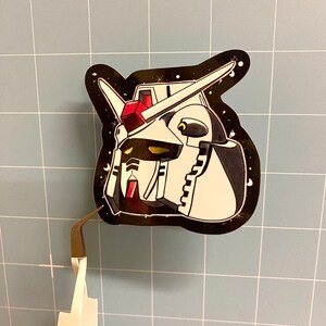 Puede incluir: Una pegatina en blanco y negro de una cabeza de robot con detalles rojos y amarillos. El robot est&aacute; sobre un fondo negro con estrellas blancas.