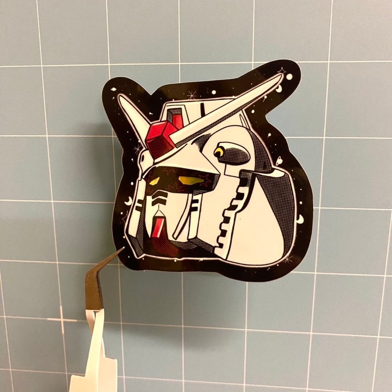 Puede incluir: Una pegatina en blanco y negro de una cabeza de robot con detalles rojos y amarillos. El robot est&aacute; sobre un fondo negro con estrellas blancas.